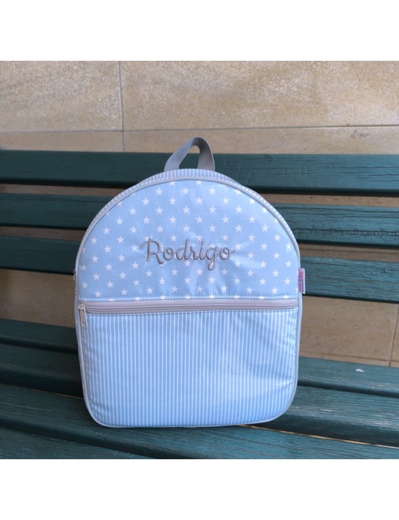 Mochila de criança azul clara com padrão de estrelas brancas conjugadas com riscas azuis e brancas. Nome Rodrigo bordado a cinzento na parte de cima da mochila. Fecho cinzento que separa a parte de cima da mochila da parte de baixo
