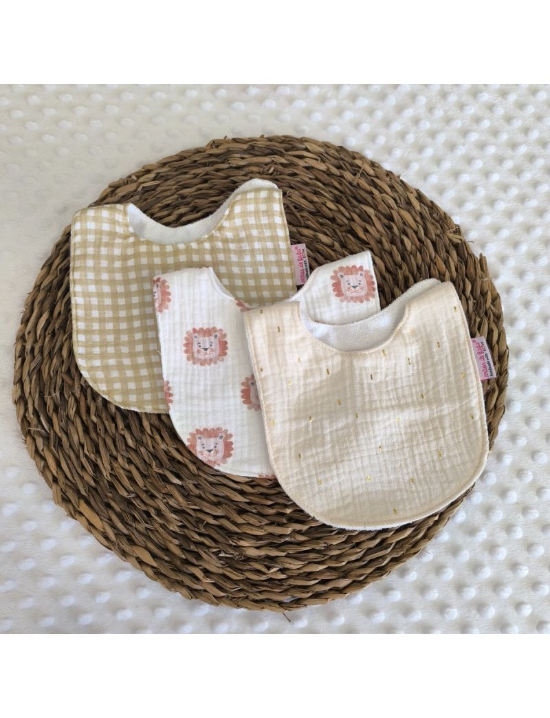 Conjunto de tr&ecirc;s babetes de mola em musselina. &Agrave; frente, um babete creme com pequenos pontos dourados; ao centro, um babete branco com ilustra&ccedil;&otilde;es de rostos de le&atilde;o; atr&aacute;s, um babete com padr&atilde;o xadrez vichy em tom mel