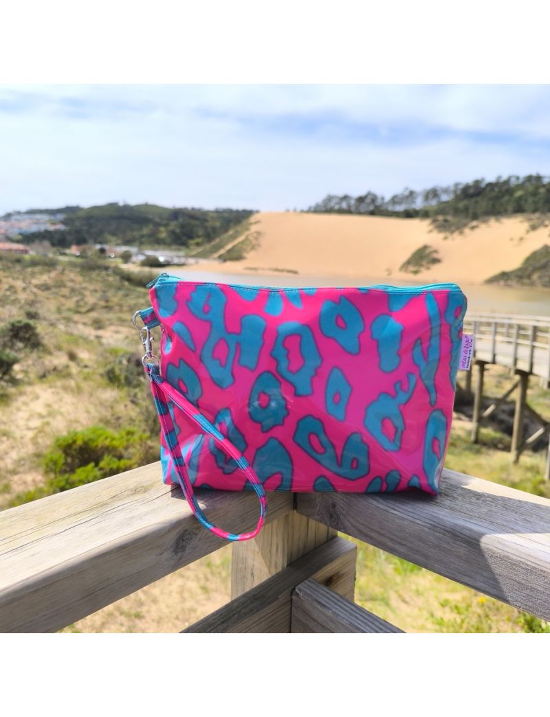 N&eacute;cessaire de design moderno com padr&atilde;o de manchas de leopardo em verde &aacute;gua escuro sobre fundo rosa neon vibrante. O material &eacute; um tecido plastificado brilhante, com fecho e al&ccedil;a de m&atilde;o condizentes. A bolsa est&aacute; pousada sobre um corrim&atilde;o de madeira r&uacute;sti