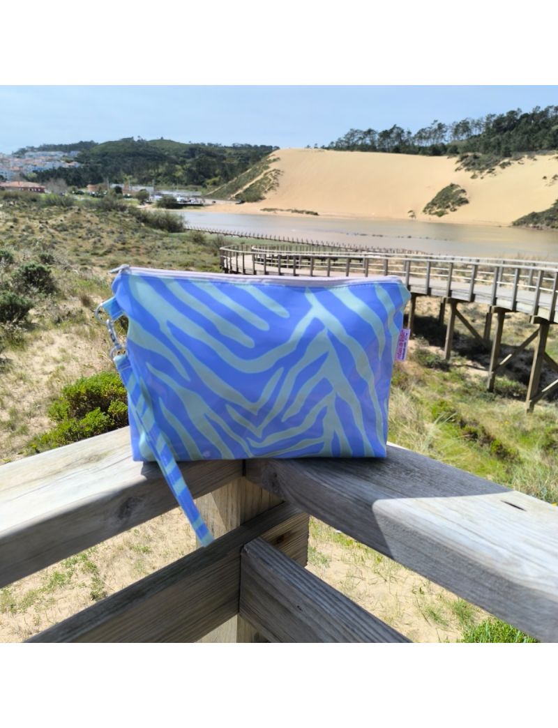 N&eacute;cessaire com padr&atilde;o zebra em tons de roxo e verde, fabricado em material plastificado brilhante. A bolsa inclui uma al&ccedil;a de m&atilde;o no mesmo padr&atilde;o e est&aacute; pousada num corrim&atilde;o de madeira de um passadi&ccedil;o