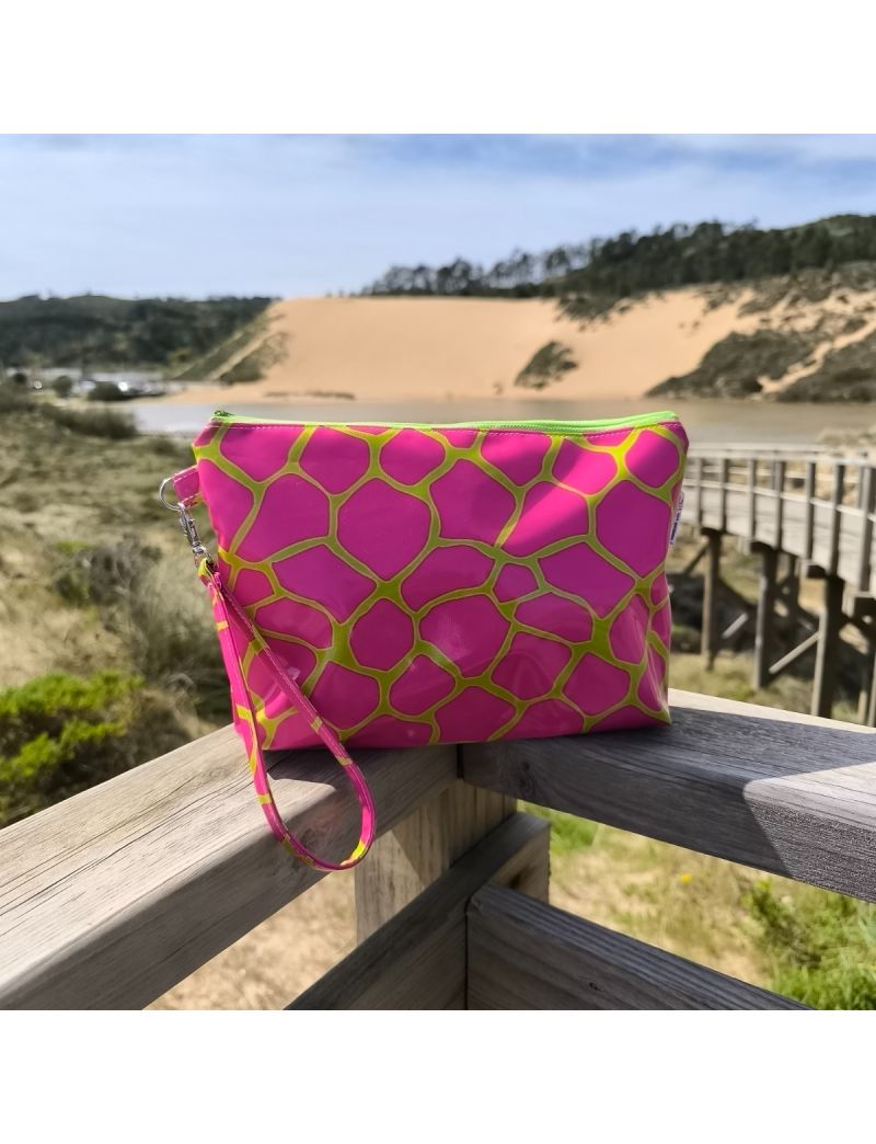 N&eacute;cessaire de design vibrante com padr&atilde;o de mosaico em rosa choque e linhas verde lima neon. A bolsa &eacute; feita de material plastificado com brilho e possui uma al&ccedil;a de m&atilde;o lateral. Est&aacute; colocada sobre um corrim&atilde;o de madeira, tendo como plano de fundo uma pa