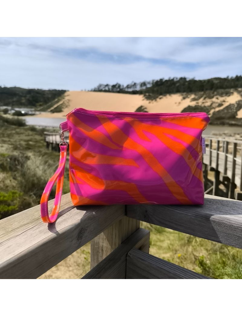 N&eacute;cessaire com padr&atilde;o zebra em tons de rosa e laranja neon, com acabamento brilhante em tecido plastificado. A bolsa possui um fecho rosa no topo e uma al&ccedil;a de m&atilde;o combinada. Est&aacute; pousada num corrim&atilde;o de madeira com uma paisagem natural de dunas