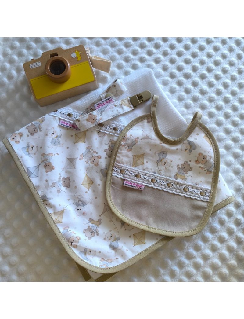 Foto de produto de enxoval de beb&eacute; com o tema ursinhos e papagaios de papel em tons de bege. O conjunto inclui uma fralda, um babete imperme&aacute;vel e uma fita de chucha com mola.
